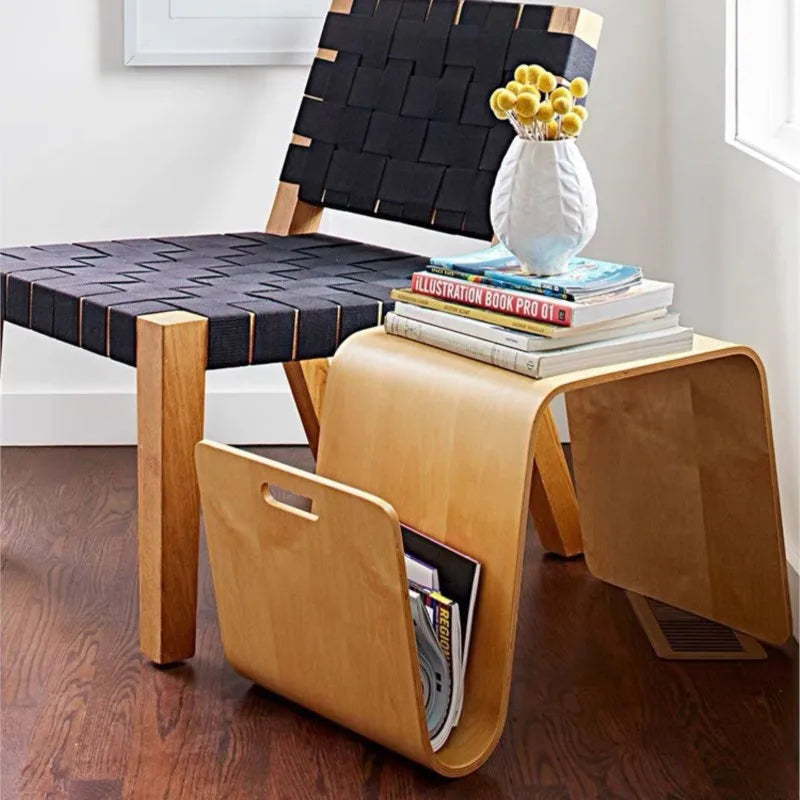 A light wood bent plywood side table with books and a vase of yellow flowers on top, and magazines stored in its curved base, next to a chair with a woven black seat.