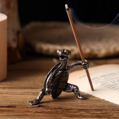 A detailed bronze incense holder shaped like a turtle holding a burning incense stick, with smoke rising, on a wooden surface.
