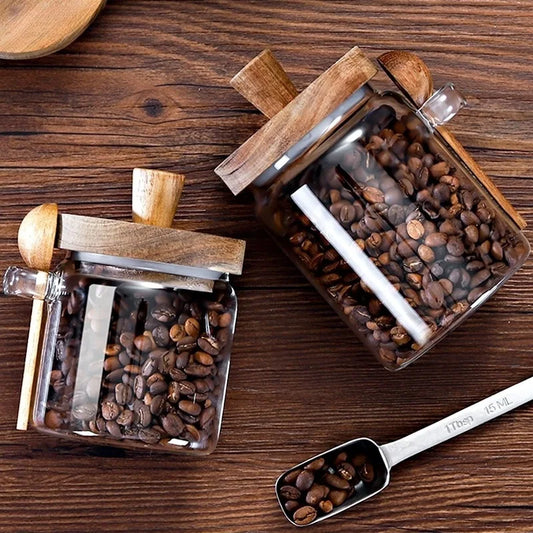 Two square glass canisters filled with coffee beans, featuring wooden lids and small wooden scoops, alongside a metal measuring spoon.