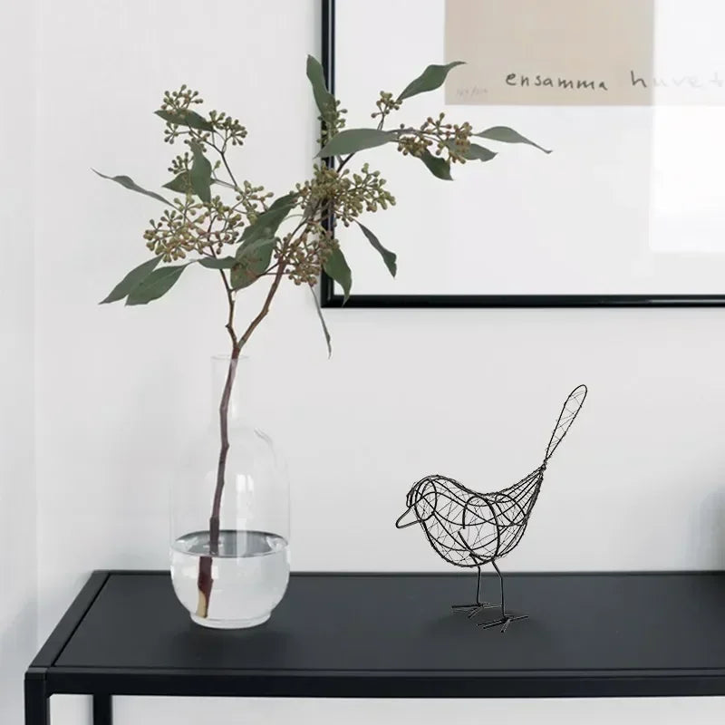 Decorative wire bird sculpture next to a clear vase with greenery on a black surface.