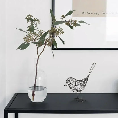 Decorative wire bird sculpture next to a clear vase with greenery on a black surface.