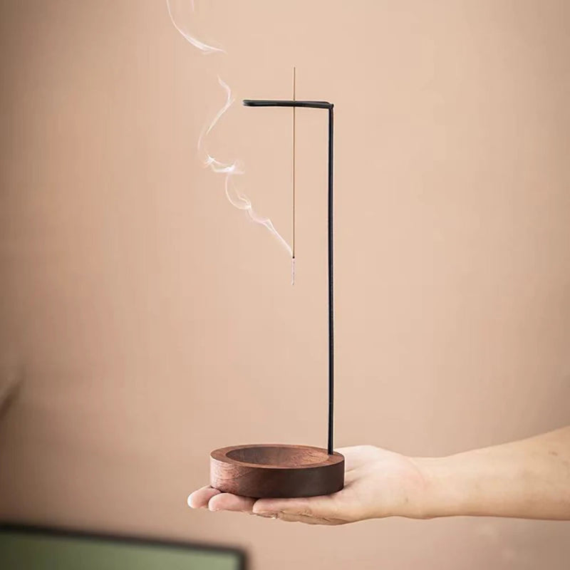 A hand holds a burning incense stick in a modern wooden and metal holder.