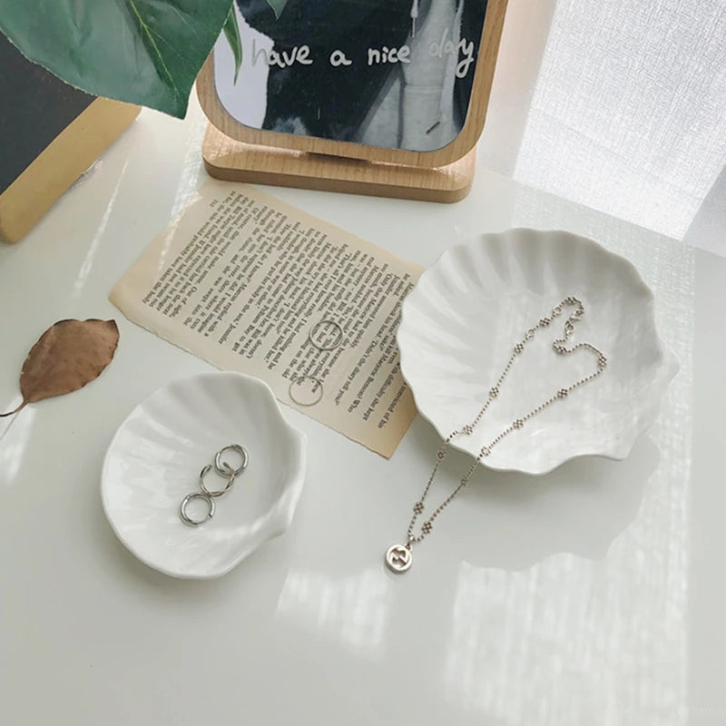 Two small, white, shell-shaped ceramic jewelry dishes, one holding three silver rings and the other holding a delicate silver necklace, resting on a white surface next to a vintage piece of paper and a mirrored sign that says "have a nice day."