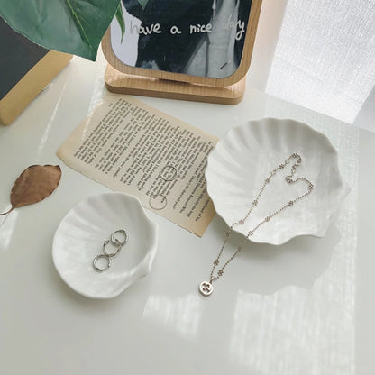 Two small, white, shell-shaped ceramic jewelry dishes, one holding three silver rings and the other holding a delicate silver necklace, resting on a white surface next to a vintage piece of paper and a mirrored sign that says "have a nice day."