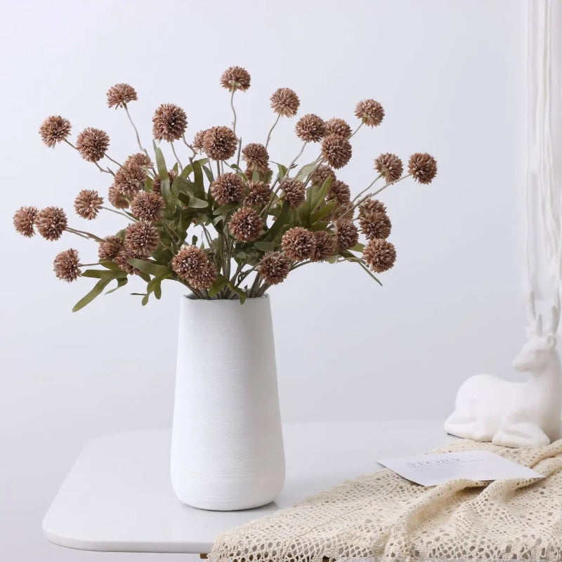 A white vase with a bouquet of brown artificial flowers and green leaves sits on a white table next to a white deer figurine and a lace cloth.