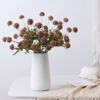 A white vase with a bouquet of brown artificial flowers and green leaves sits on a white table next to a white deer figurine and a lace cloth.