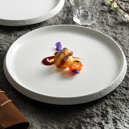 White porcelain plate with a textured stone-like edge, artfully plated with a gourmet dish, set on a textured surface with elegant tableware.