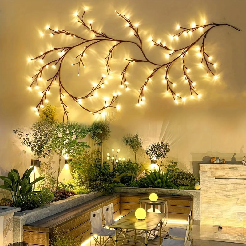 A lit string of branch-shaped lights hangs on a wall above a modern outdoor patio with a dining table and various plants.