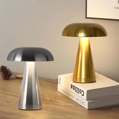 Two mushroom-shaped lamps, one silver and one gold, are displayed on a wooden surface, with the gold lamp resting on two stacked books titled "DECOR ALL-AMERICAN FLAIR Collins," while a pinecone and small wooden disc lie beside the silver lamp, all set against a blurry background featuring a framed artwork with the text "TIM" partially visible.