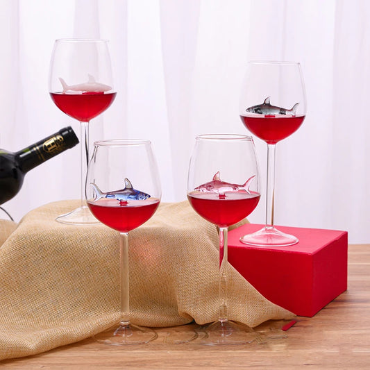 A set of four clear wine glasses, each containing a small, colored glass shark figurine suspended inside, filled with a red liquid.