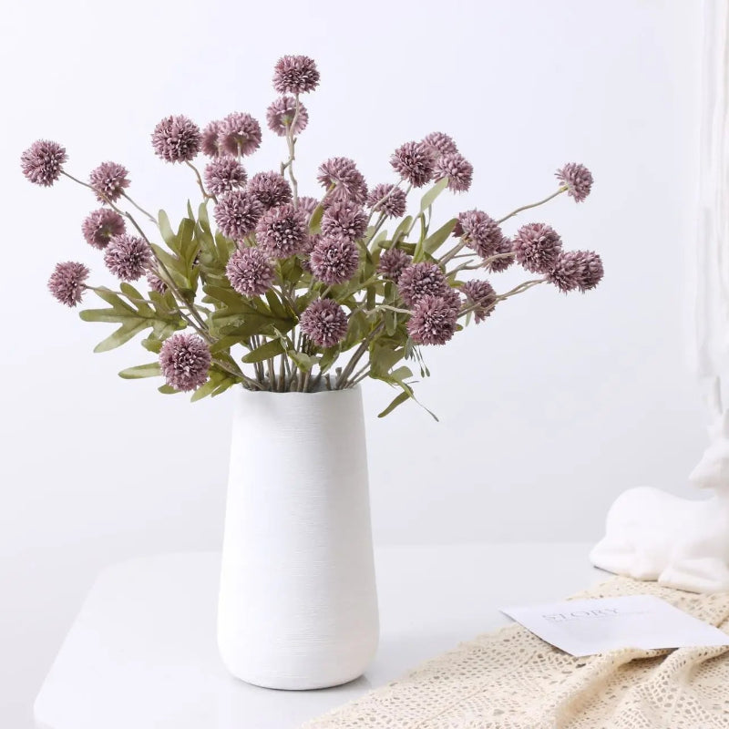 A white vase with purple artificial flowers and green leaves sits on a white table next to a white deer figurine and a lace cloth.