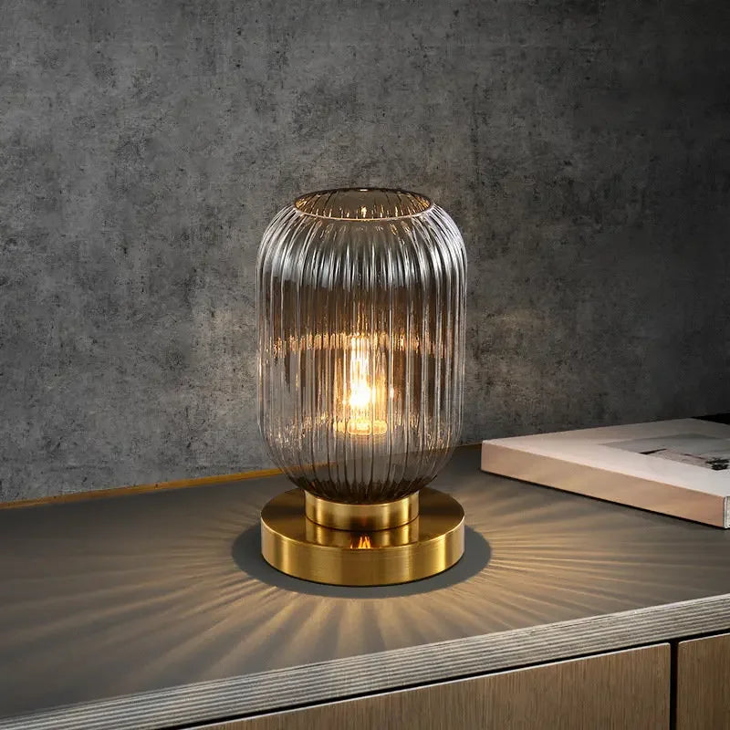A lit, ribbed, smoky gray glass table lamp with a gold base sits on a dark gray surface, casting light patterns. A stack of papers rests behind it against a textured gray wall.
