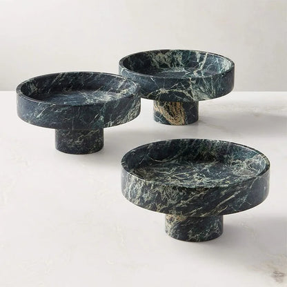 Three round, dark green marble pedestal stands with a polished surface and unique white veining patterns introduce a modern twist. They sit elegantly on a pale, smooth surface against a neutral background, perfect for displaying a premium fruit bowl.