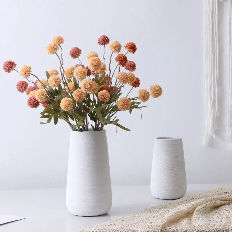 A tall white vase with artificial peach and rust-colored flowers stands next to a shorter white vase and a lace throw.