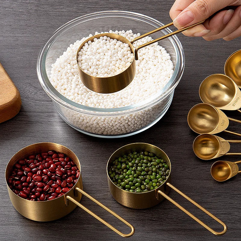 A set of gold-colored measuring cups filled with various beans and grains, with one cup being held over a large glass bowl.