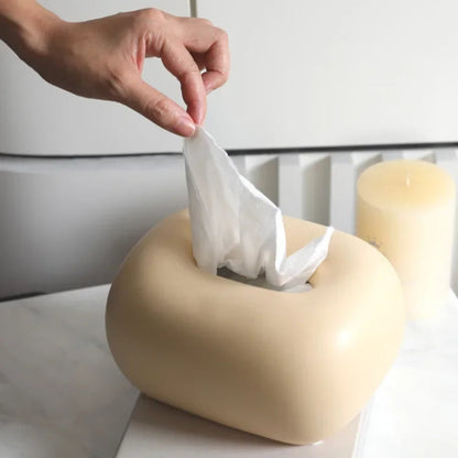 A hand pulls a tissue from a donut-shaped, light yellow tissue box holder that sits on a white marble surface.