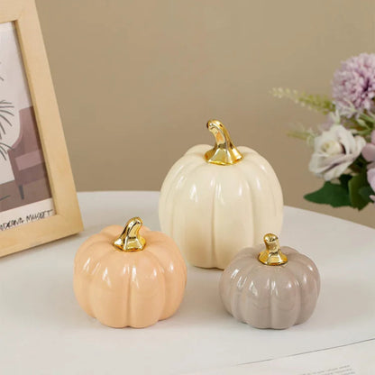 Three ceramic pumpkins, in cream, peach, and gray, with gold stems, are arranged on a white table next to a framed picture and a blurred floral arrangement.