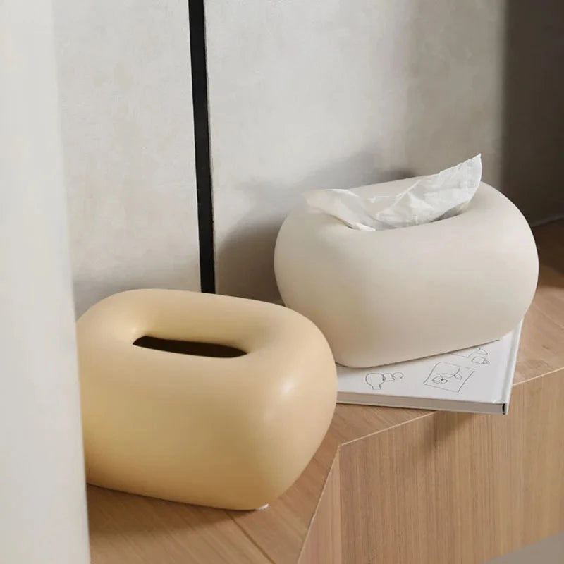 Two donut-shaped tissue box holders, one light yellow and one light beige, sit on a light wood surface.