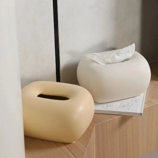 Two donut-shaped tissue box holders, one light yellow and one light beige, sit on a light wood surface.