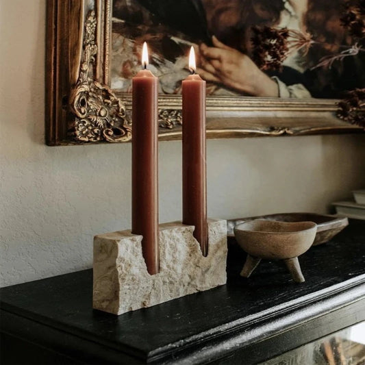 Two lit, dark red taper candles in a textured, light-colored travertine stone block holder, sitting on a dark surface with a small, light-colored bowl, against a wall with a framed picture.