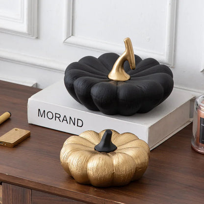 A small, textured gold pumpkin with a black stem sits on a wooden surface in front of a larger, textured black pumpkin with a gold stem.