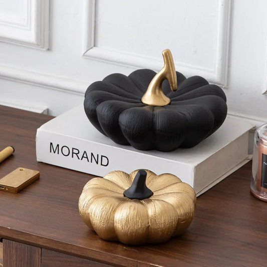 A small, textured gold pumpkin with a black stem sits on a wooden surface in front of a larger, textured black pumpkin with a gold stem.