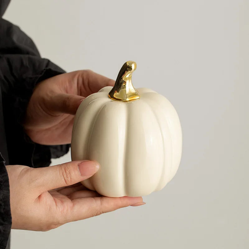 A person holds a small, cream-colored ceramic pumpkin with a gold stem in their hands.