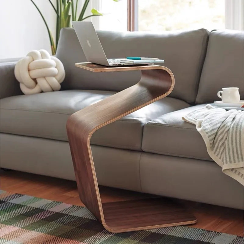 A wooden, Z-shaped side table with a laptop and pen on top, placed next to a gray sofa with a knot pillow and a white mug.
