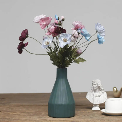 A dark gray faceted vase holds a bouquet of multicolored poppies, next to a white bust and a teacup.