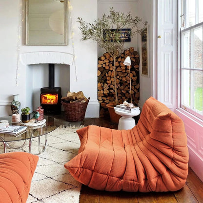 A cozy living room featuring an orange Togo armchair and footrest, a lit fireplace, and a stack of firewood.