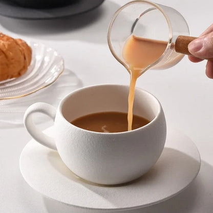 A hand pouring liquid from a glass creamer into a white textured coffee cup.