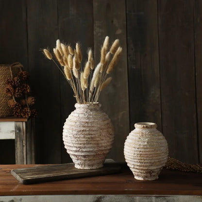 Two textured vases with dried plants on a wooden surface against a dark wooden wall.