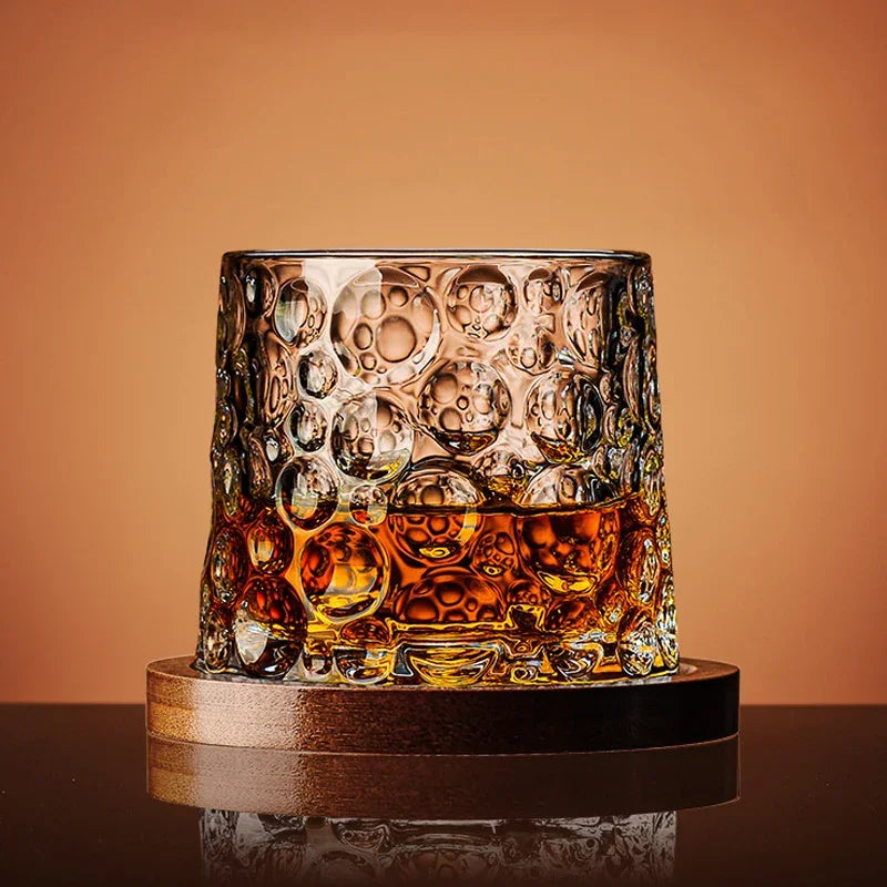 A round, textured whiskey glass filled with amber liquid sits on a wooden coaster.