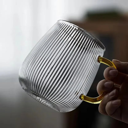 A hand holding a clear, empty, ribbed glass mug with a gold-colored handle.