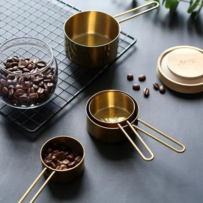 A brass measuring cup set, with one cup filled with coffee beans, sits on a black wire rack next to a glass jar of coffee beans and a lidded container.