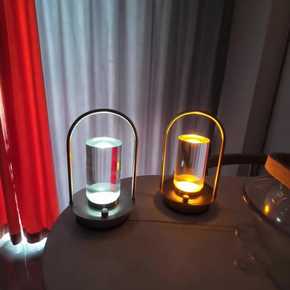 Two matching portable table lamps with cylindrical glass shades and metal frames, one unlit and one glowing with a warm light, sitting side-by-side on a table.