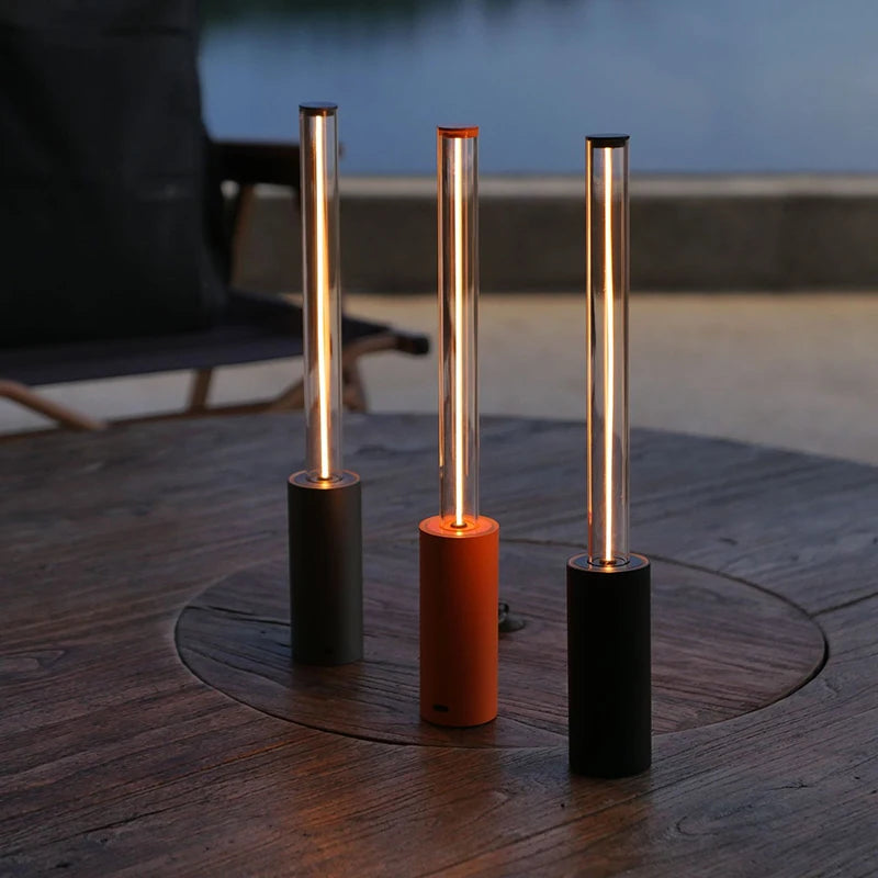 Three small, cylindrical lamps with glowing tubes, one gray, one orange, and one black, on a round wooden table outdoors.