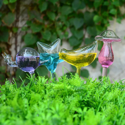 Four clear glass plant waterers, shaped like a snail, butterfly, bird, and mushroom, are inserted into a patch of vibrant green foliage.