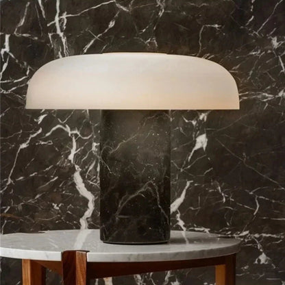 A modern mushroom-shaped table lamp with a dark marble base and a white dome shade, on a white marble table with a dark, veined marble wall behind it.