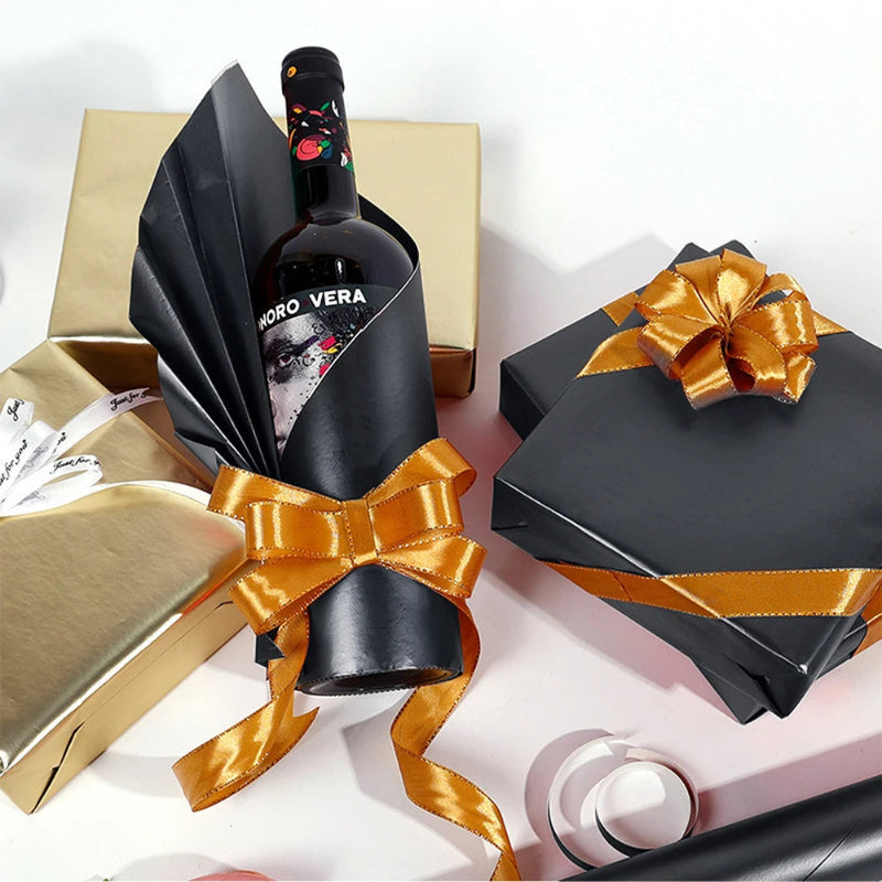 Bottle of wine wrapped in black paper with a gold ribbon, surrounded by gift boxes on a white background.