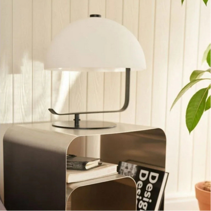 A half-dome shaped lamp with a black metal base and white shade sits on a silver, bent metal nightstand with rounded corners.
