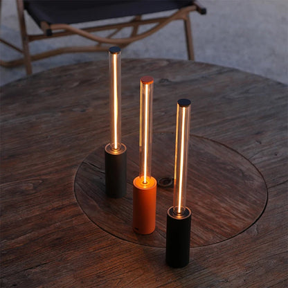 Three different colored long, cylindrical lamps with glowing tubes sitting on a round wooden table.