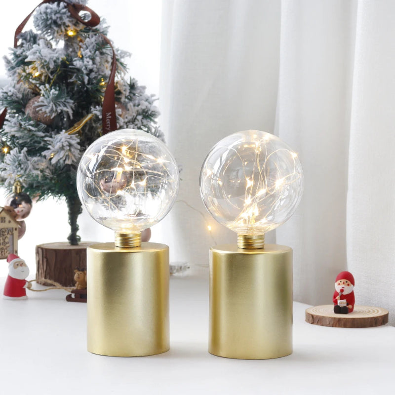 A pair of modern table lamps with gold cylindrical bases and clear glass globe shades filled with warm white fairy lights, with a small decorated Christmas tree and figurines in the background.