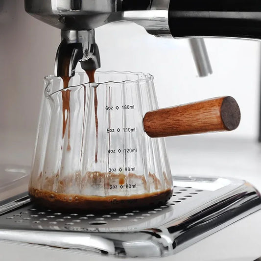 Clear glass measuring cup with a ribbed design and wooden handle, placed under an espresso machine as fresh espresso pours in. Measurement markings in ounces and milliliters are visible on the cup.