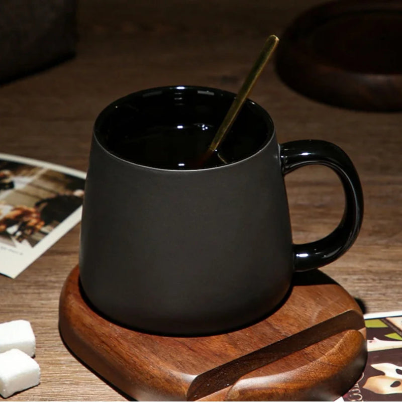 A black mug with a gold spoon on a wooden coaster, with sugar cubes and old photographs nearby.