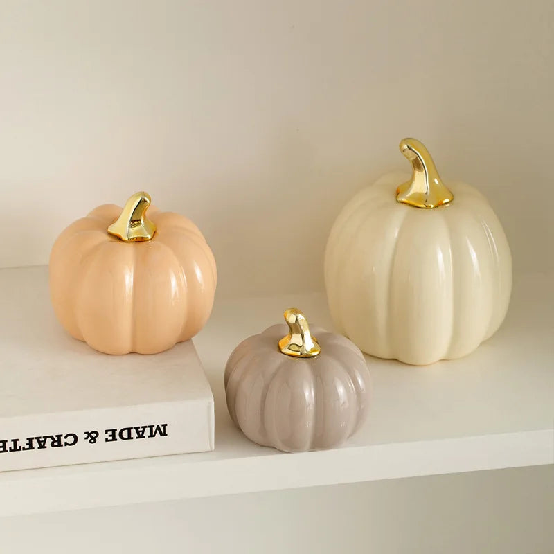 Three ceramic pumpkins, in peach, gray, and cream, with gold stems, sit on a white shelf next to a book.