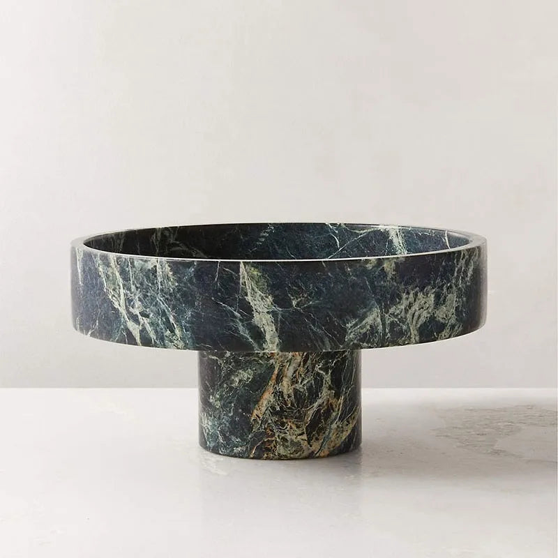 A round, elevated green marble bowl with a cylindrical base, featuring white and brown veining, adds a modern twist to any decor. This premium fruit bowl sits on a smooth, light-colored surface against an off-white wall.