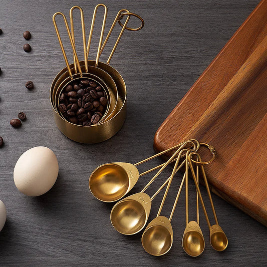 A set of nested, gold-colored measuring cups and spoons on a dark surface, with coffee beans in the largest cup and loose coffee beans and eggs nearby.