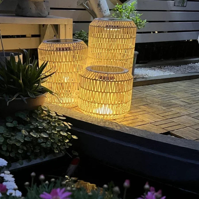 Woven outdoor lanterns emitting a warm glow in a garden setting with plants and flowers.