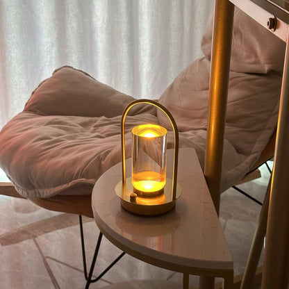 A lit, portable table lamp with a gold metal frame and a glass shade, placed on a small, half-moon shaped side table next to a rocking chair with a light-colored cushion.